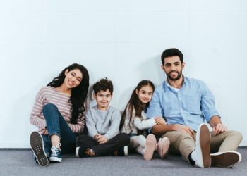 cheerful hispanic family smiling while sitting on floor near white wall