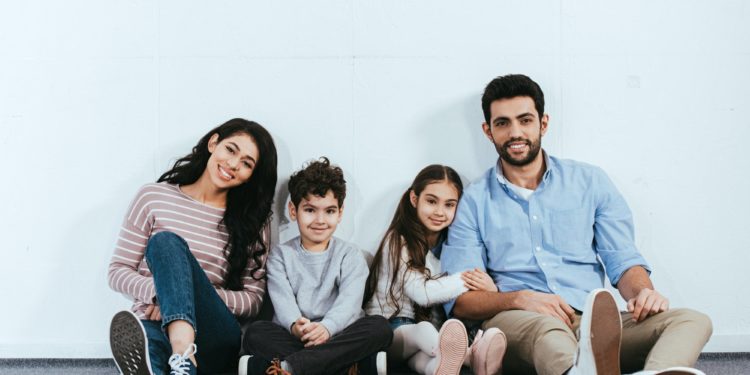 cheerful hispanic family smiling while sitting on floor near white wall